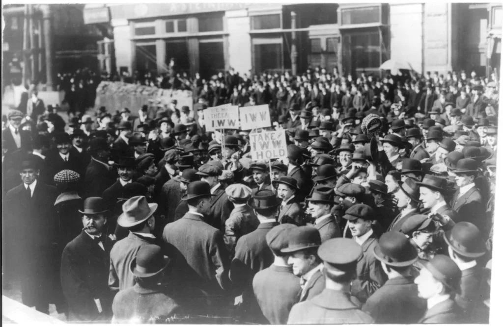 黑白老照片,1914年纽约街头聚集着密集人群,多为戴礼帽、穿西装的男性,人群中举着写有“IWW”字样的标语牌。