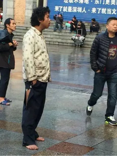雨后广场上,一名赤脚、衣着邋遢的男子独自站立,背后横幅写着“来了就是深圳人”,周围有行人经过,台阶上坐着几个人。