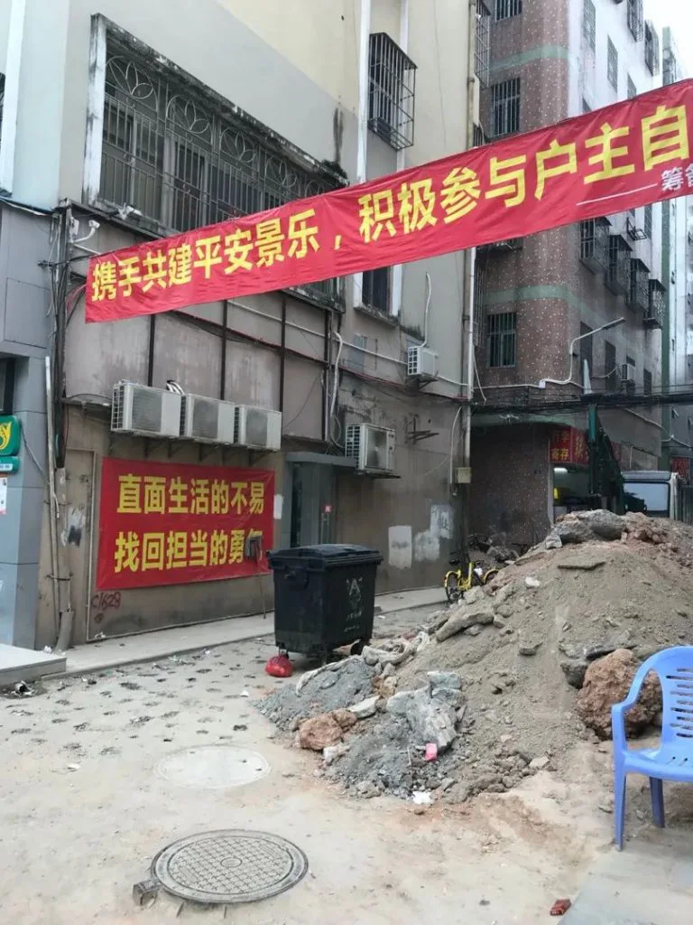 城中村巷道内,头顶悬挂红底黄字横幅“携手共建平安景乐,积极参与户主自……”,墙上另贴有图注所引标语,路旁堆放建筑渣土、垃圾桶和一把蓝色塑料椅。