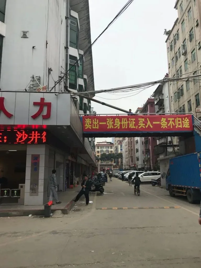 街道上方悬挂红底黄字横幅“卖出一张身份证,买入一条不归途”,横幅下方是写着“人力”的招牌店铺,街上有行人、电动车和一辆蓝色卡车经过。