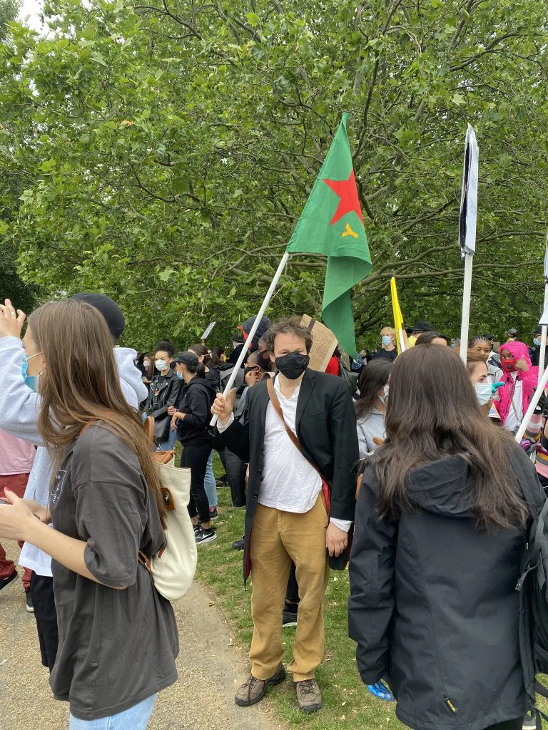 格雷伯戴口罩站在户外人群中,手举一面绿底红星旗帜,周围是同样戴口罩的抗议者,背景为树木环绕的公园。