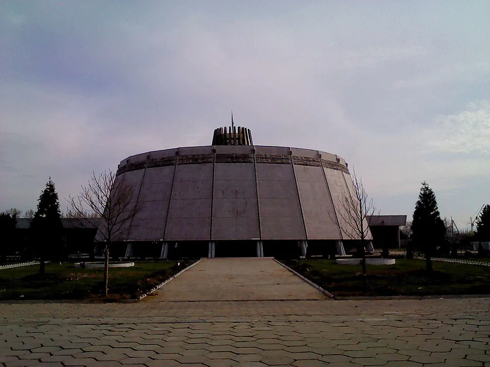 一座鼓形混凝土建筑架于立柱之上,顶部有金属装饰与旗杆,前方铺石步道两侧种着光秃的树木。