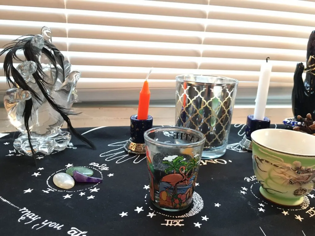 祭坛台面铺着印有星月符号的黑布，摆放一支橙色蜡烛、一尊黑发玻璃女巫摆件、几只印花玻璃杯和一只龙纹瓷碗，桌上还散落着贝壳与松果。