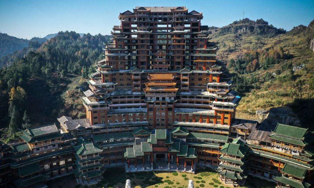 航拍视角下的一座巨型仿古建筑群,主楼呈金字塔状层层收窄至顶部,四周环绕绿色琉璃瓦的低矮附属建筑,坐落于群山环抱的谷地中。