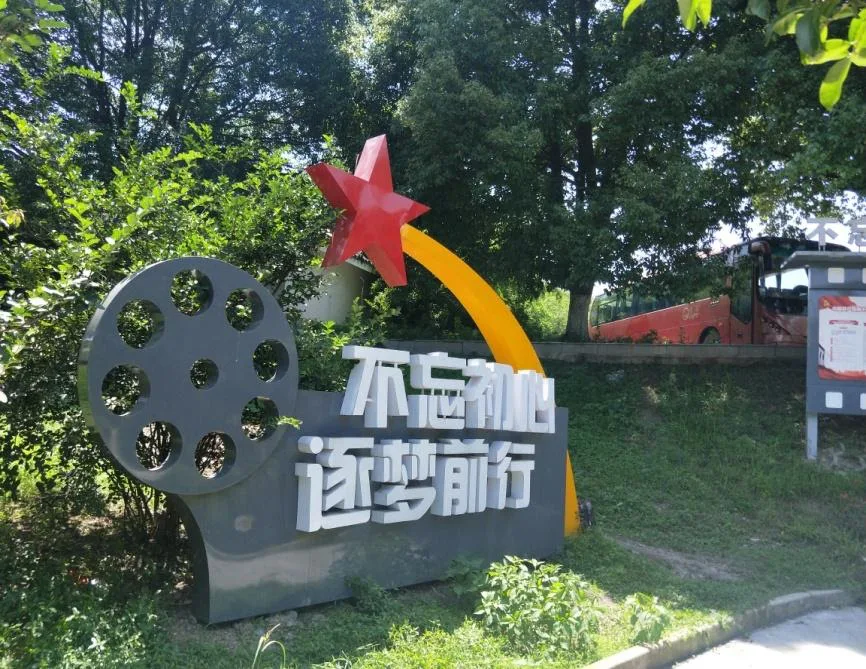 路边一座雕塑标语牌,灰色胶片卷轴造型旁立着红色五角星与黄色弧线,下方白色大字写着'不忘初心 逐梦前行',背景是树丛和一辆停靠的大巴。
