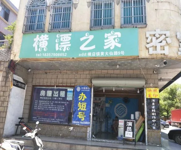 临街店铺,绿色招牌上写着'横漂之家'及电话、地址,楼下橱窗贴着'房屋出租''办短号''演员'等告示,门前停着一辆电动车。