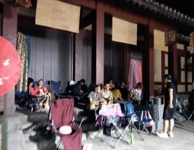 夜晚,一群人坐在折叠椅上,在仿古木结构建筑的檐廊下休息,旁边挂着戏服和红灯笼。