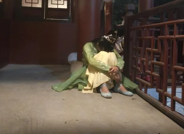 夜间,一名头戴发饰的女性蜷坐在木质回廊地面上休息,身穿绿黄相间的古装长裙,双手环抱膝盖低着头。