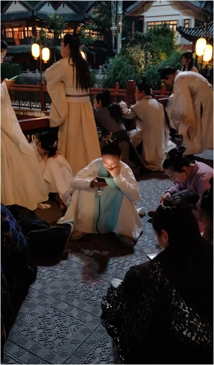 夜晚,多名身着古装的群演或坐或蹲在铺着花纹地砖的场地上休息,其中一人低头看着手机,背景是亮灯的仿古建筑。