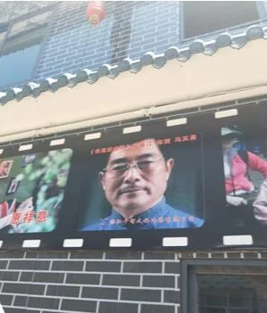 民居外墙上挂着多幅长方形海报,居中一幅是戴眼镜男子的正装肖像照,两侧海报部分被遮挡,隐约可见身着古装的人物。