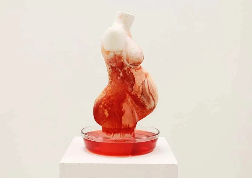一尊怀孕女性躯干雕塑立于白色展台的透明浅盘中,雕塑表层渗着暗红色痕迹,从上半身向下流淌汇入盘中鲜红的液体,如同渗血或熔化。