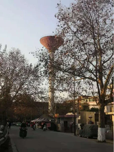 一座高耸的漏斗形水塔矗立在光秃秃的行道树之间，塔身细长、顶部呈伞状展开，背景是老旧居民楼，塔下街道上有行人和摩托车经过。