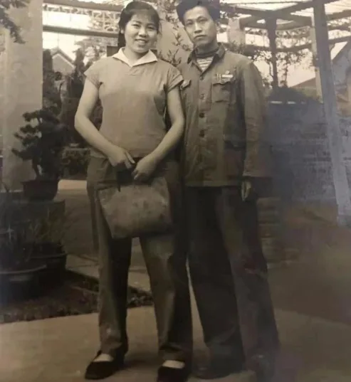 一张黑白老照片，年轻女子留短发、穿短袖衬衫和长裤，手提布包，身旁是穿深色制服式外套的年轻男子，两人站在藤架下，一旁摆着盆景。