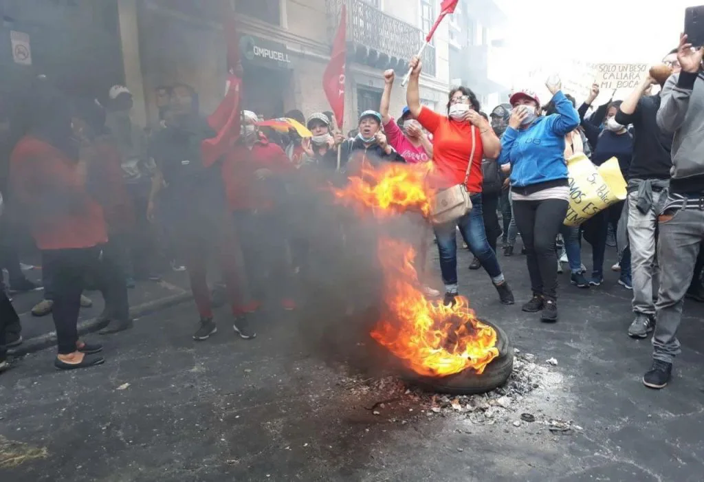 街头一只轮胎燃烧冒出浓烟，周围抗议者举拳呐喊，有人挥舞红旗、举着标语牌，多数人戴口罩。