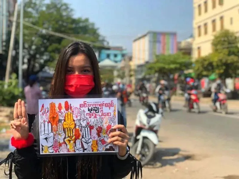 一位年轻女性戴红色口罩,比出三指抗议手势,双手举着印有缅甸文标语和多彩举手图案的传单,身后是仰光街头往来的摩托车和行人。