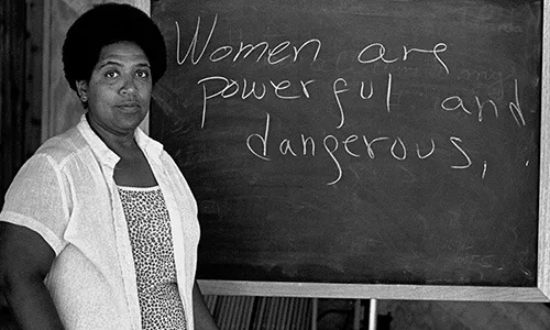 Audre Lorde站在黑板前,黑板上手写着“Women are powerful and dangerous,”(女人是强大而危险的)。