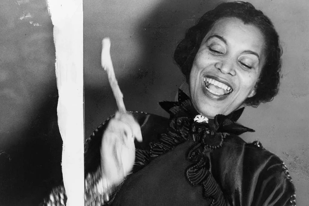 Zora Neale Hurston黑白照片,她开怀大笑,抬手做出动作,身穿带蕾丝立领的深色礼服。