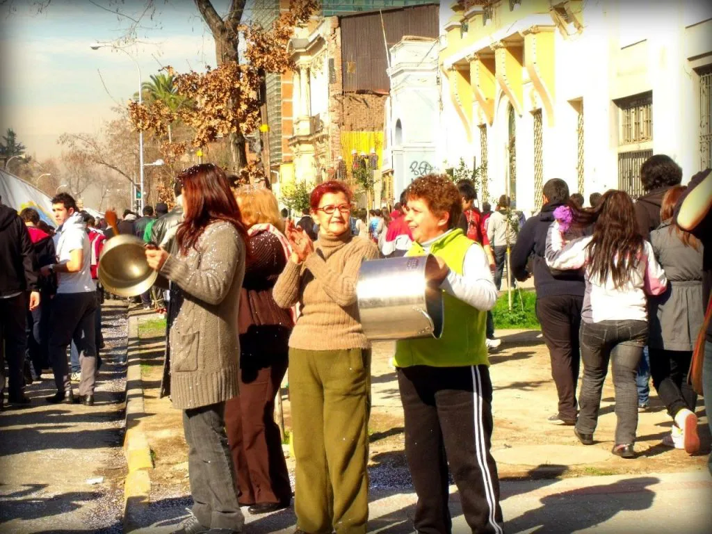 街头人群列队行进,前景两位年长女性一人举着长柄平底锅、一人抱着金属锅相互敲击,两侧是欧式风格建筑,阳光下街道人潮涌动。