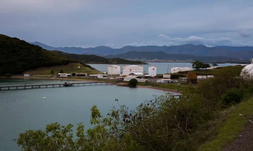 海湾旁的石油仓储罐区，几座白色圆形油罐立在岸边，背后是连绵山脉，海面平静，前景为绿色灌木与山坡。