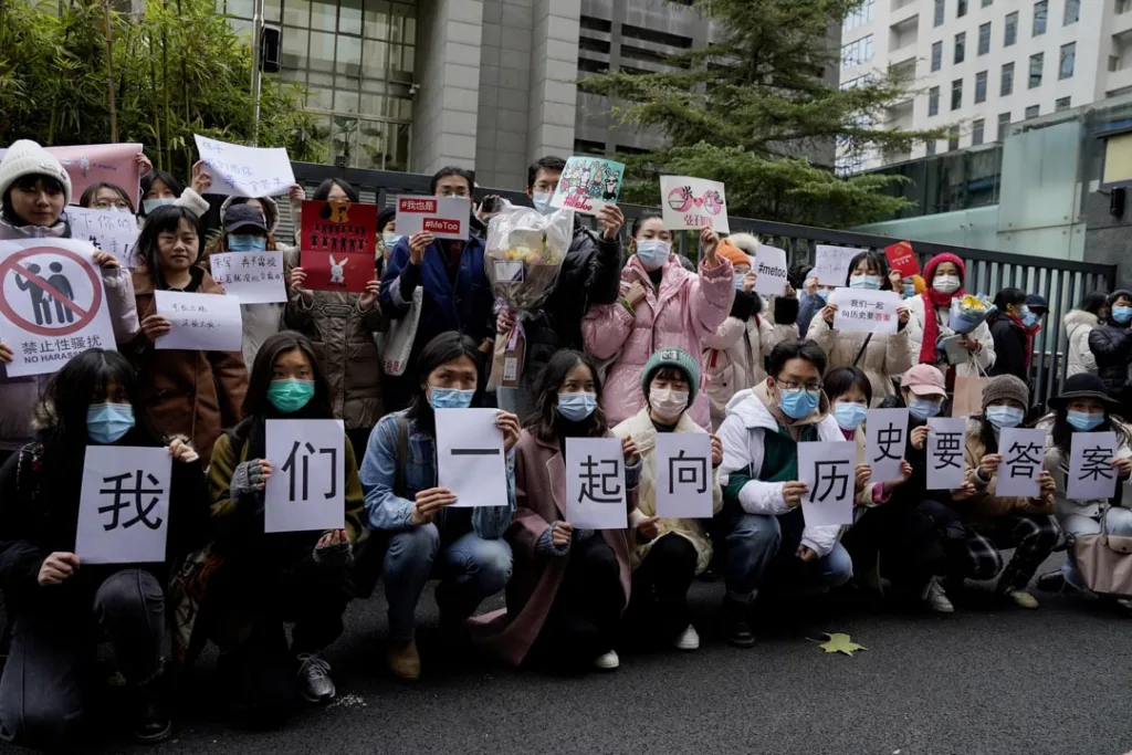 一群戴口罩的人聚集在建筑外声援,前排举牌拼出“我们一起向历史要答案”,其他人举着写有#MeToo、“禁止性骚扰”等字样的标语和鲜花。