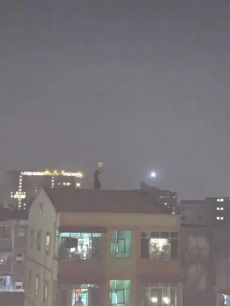 夜晚楼顶剪影:一人站在天台边缘,双手举起圆盘状金属容器对着夜空中的圆月,远处高楼灯火与工地照明灯可见。