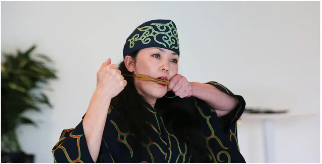 身着深色阿伊努族传统长袍与头巾的女性，衣物绣有绿色漩涡纹样，双手举着竹制口簧琴凑近嘴边演奏，室内背景可见绿植。