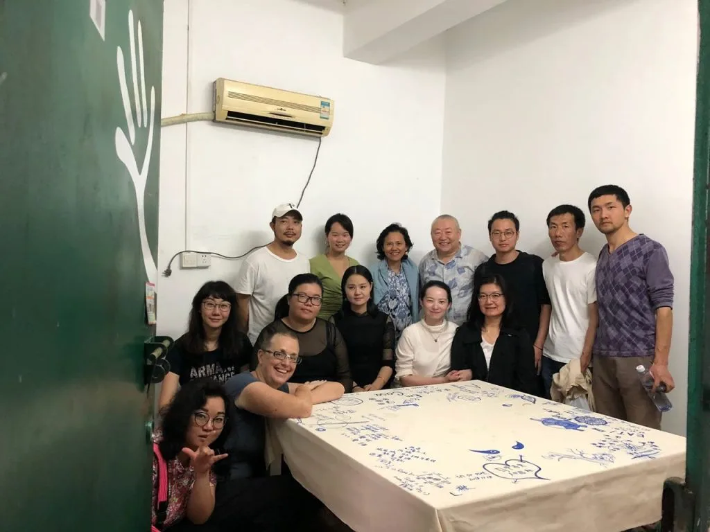 十余人在室内合影,围站在一张铺着白布的长桌旁,白布上绘满蓝色手绘图案与文字,一旁绿色房门上印着白色手掌图案。