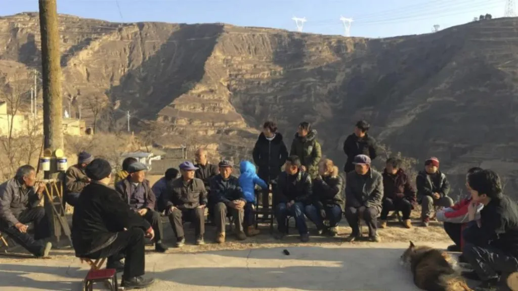 一群村民坐在小板凳上围成半圈,在黄土沟壑地貌前露天开会,一旁有狗趴卧地面。