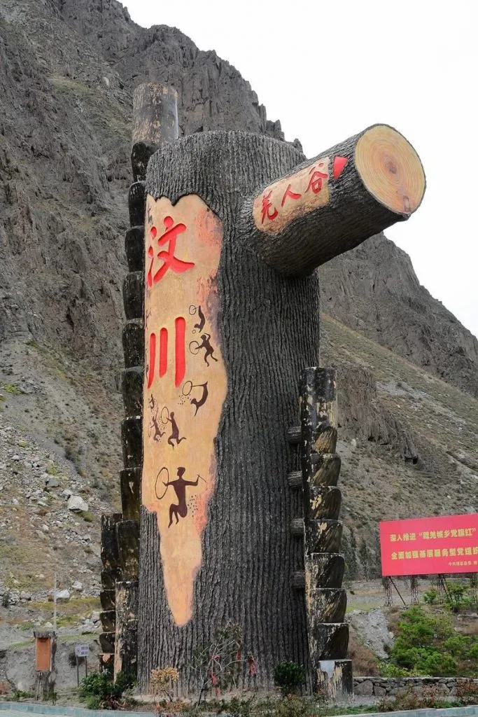 一座仿树桩造型的巨型雕塑矗立在山前,正面用红色大字写着“汶川”,上面绘有类似岩画风格的人物与动物图案,右侧断枝造型上写着“尧人谷”,背景是陡峭的山体。