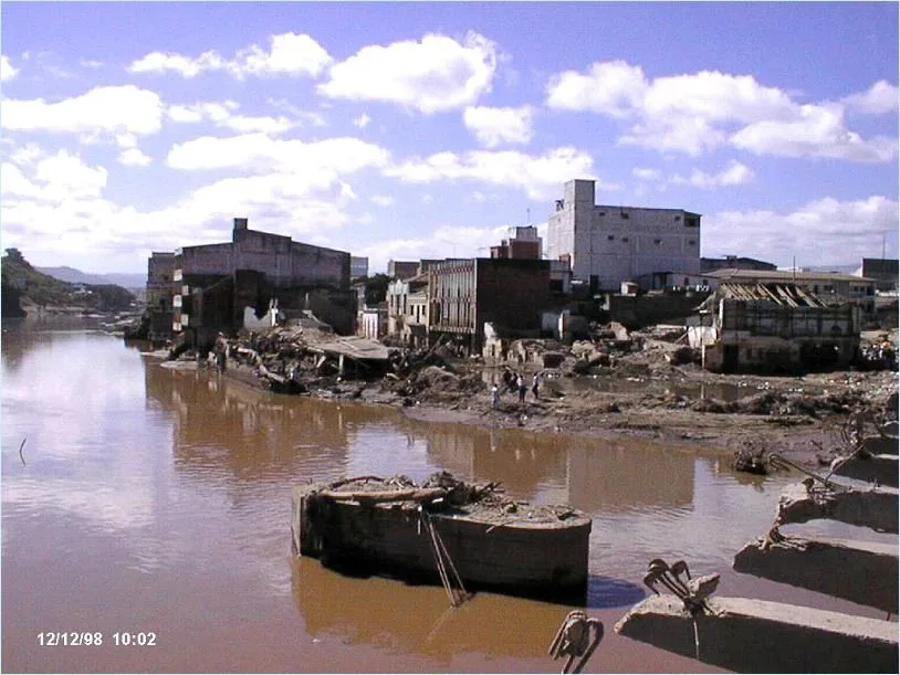 浑浊的洪水淹没河岸城区,两侧建筑严重损毁、墙体倒塌,几名人员在泥泞中查看,画面左下角有日期时间戳“12/12/98 10:02”。
