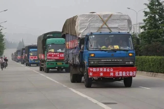 一列满载物资的卡车车队行驶在公路上,最前方卡车挂着红色横幅,写着“中国扶贫基金会 新浪网 抗震救灾物资”,路边有骑车人经过。