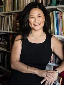 一位长发女性微笑站立,身后是摆满书籍的书架。