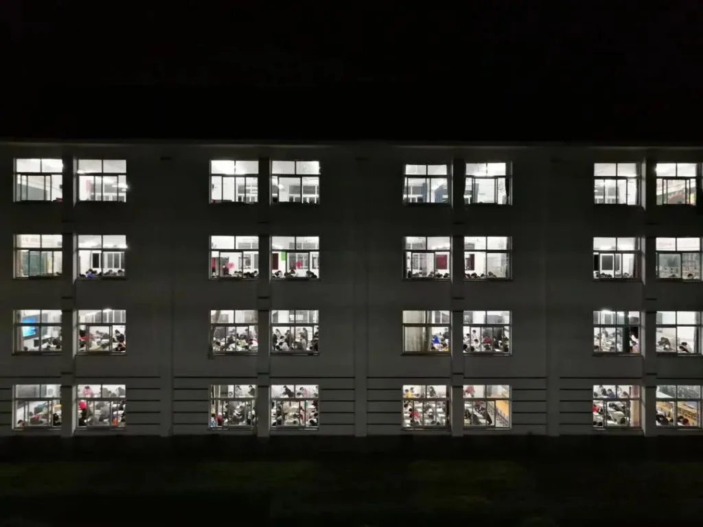 夜晚拍摄的教学楼外观,四层楼的窗户内灯火通明,可见学生伏案自习的身影。