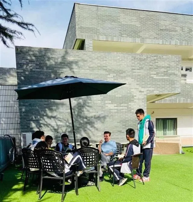 校园草坪上,几名穿校服的学生围坐在遮阳伞下的桌旁,与一位外籍男教师交谈,另一名学生站立一旁,背景是现代风格教学楼。