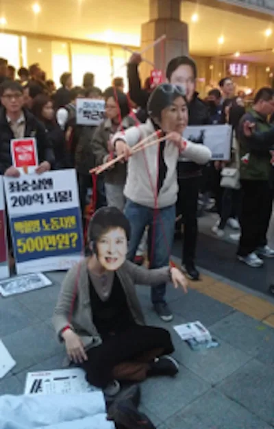 一人戴着人形面具如提线木偶般被红绳牵引倒地,周围人群举着韩文标语牌,背景是灯火通明的建筑。