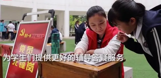一位身穿红马甲的女老师俯身在户外课桌前为学生讲解习题,周围是操场和其他学生,画面字幕写着“为学生们提供更好的辅导答疑服务”,红色展板印有“党员先锋岗”字样。