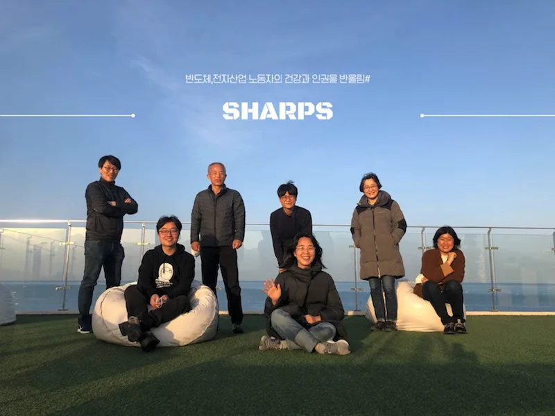 七人在铺有人工草坪的天台合影,身后是海景与蓝天,画面上方印有“SHARPS”字样。