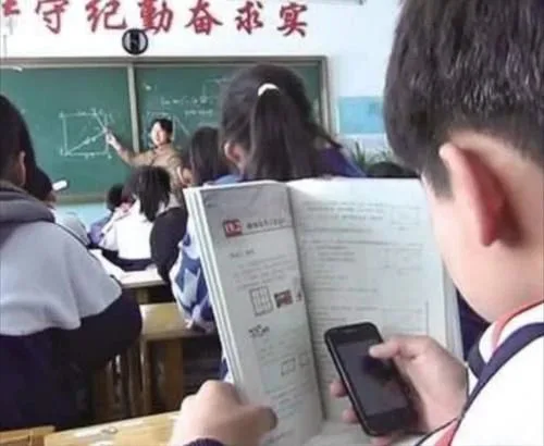 课堂上一名男生用摊开的课本挡着手机低头玩手机,讲台前老师在黑板上讲解几何图形,周围同学埋头听课