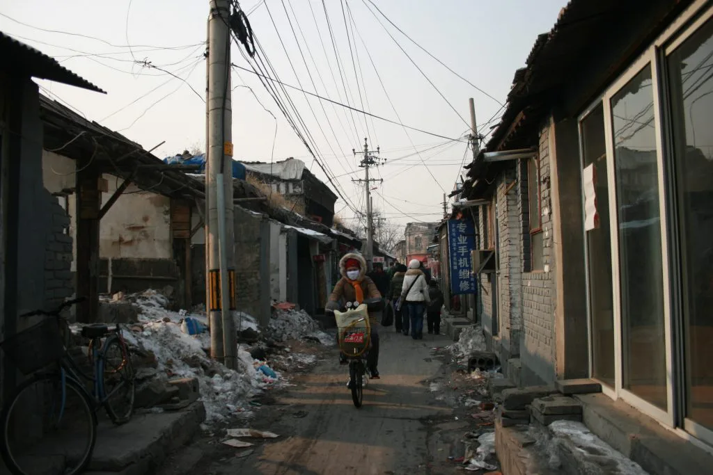 一位骑车人穿过大栅栏胡同,两侧是低矮平房,路边堆着积雪和杂物,头顶电线密布,远处几位行人在窄巷中行走。