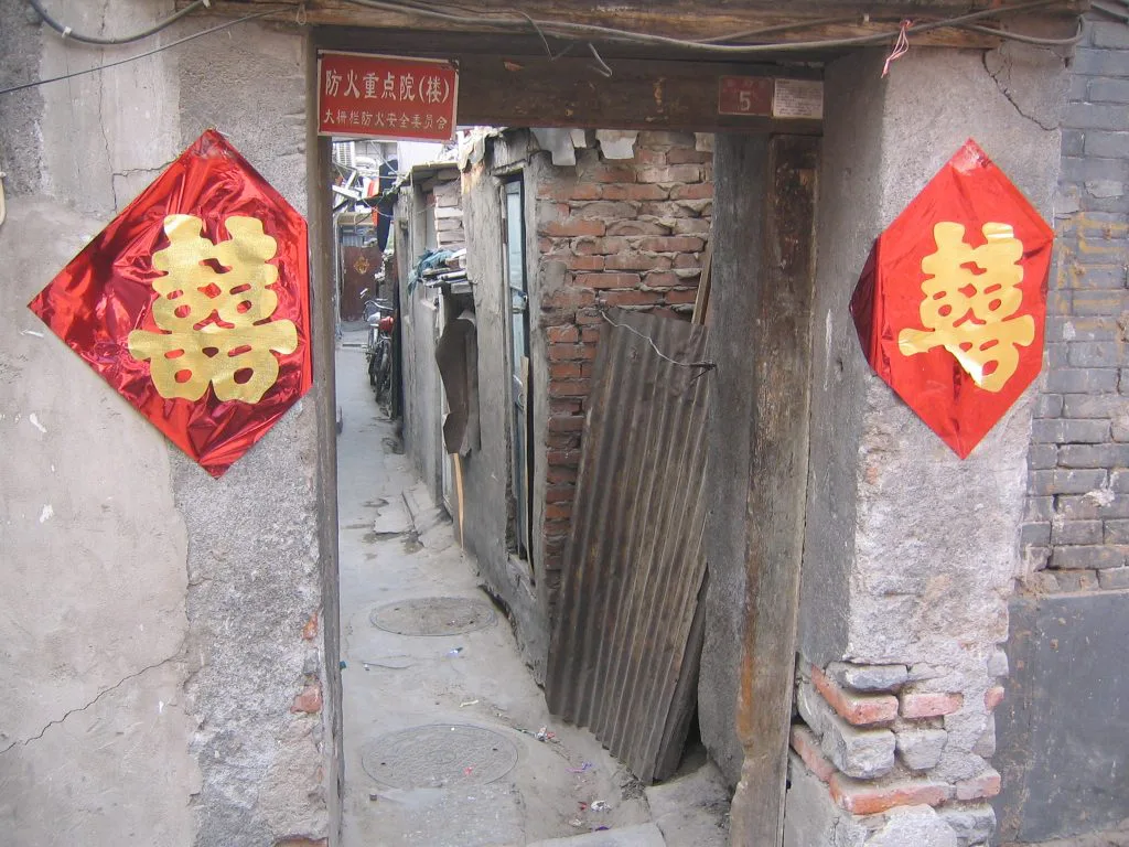 大杂院门口两侧贴着红色喜字剪纸,门框上挂着“防火重点院”标牌,门内是狭窄破旧的过道,堆放着木板等杂物。