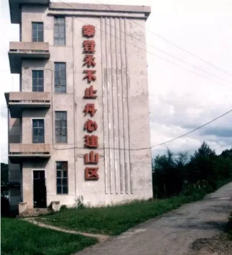 一栋灰白色多层建筑侧面,竖排红色大字写着'攀登永不止,丹心建山区',楼前是碎石路和杂草地。