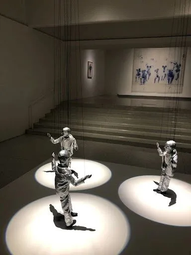 昏暗展厅中,三个身穿银色金属光泽西装的人偶站在聚光灯圆圈中,头部、手部和背后延伸出黑色细绳直达天花板,背景墙上挂着一幅人物剪影画作。
