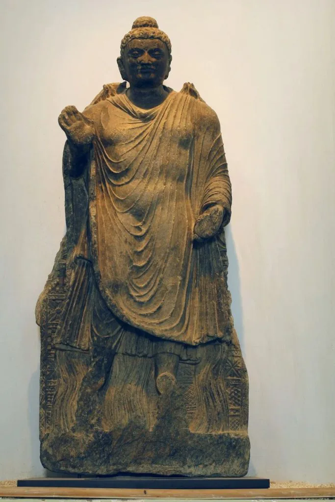 一尊立姿石雕佛像,身披褶皱厚重的长袍,一手上举做手印,另一手垂放,衣纹雕刻细腻。