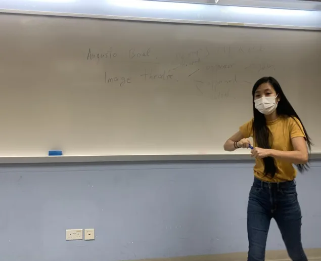 教室内,一名戴口罩、身穿黄色上衣的女子双手摆出造型动作,身后白板上手写着「Augusto Boal」「Image theatre」等字样。