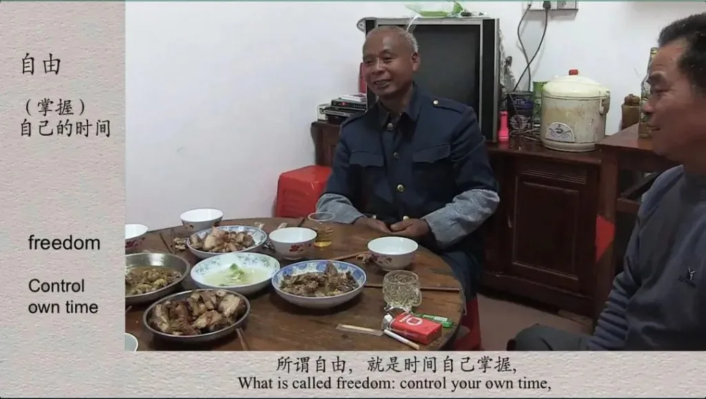 视频截图，两位男子坐在摆满家常菜肴的八仙桌旁交谈，画面左侧叠加文字“自由（掌握）自己的时间”及英文翻译，底部字幕写着“所谓自由，就是时间自己掌握”。