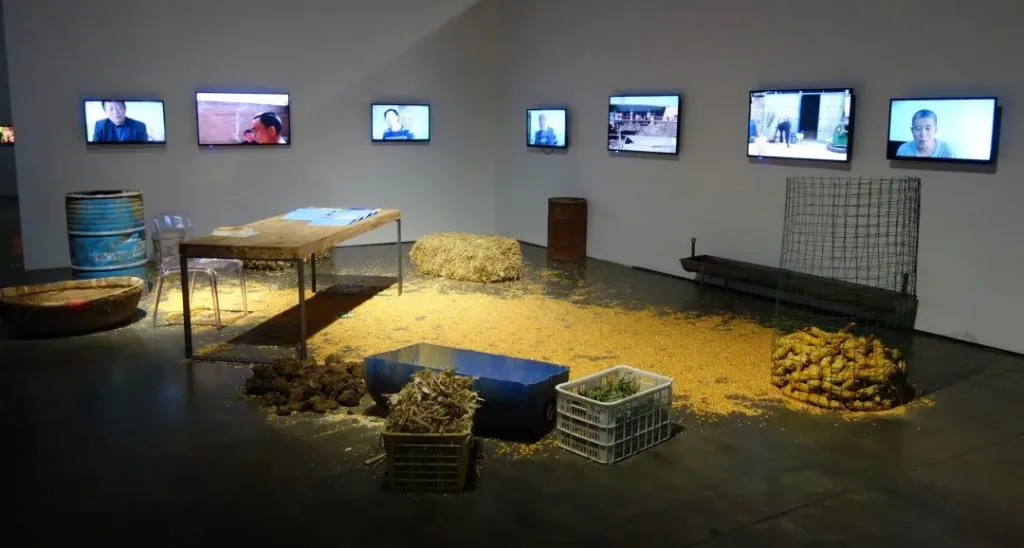 展厅一角，墙上挂着七块显示屏播放访谈影像，地面铺满玉米粒与稻草，散置着塑料筐、木箱、铁桶和一张写字桌，构成装置现场。