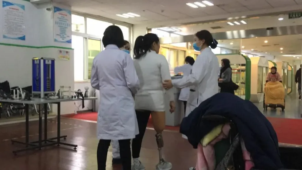 两名穿白大褂的治疗师搀扶着一名戴大腿假肢的女性在红色地垫上练习行走,大厅内可见轮椅与其他人员活动。