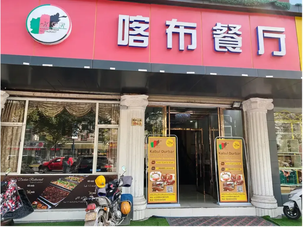 临街的阿富汗餐厅门面,招牌写着“喀布餐厅”,左侧圆形标志印有阿富汗国旗,门口立着写有“Kabul Durbar”的双语菜单牌和二维码,门前停着摩托车。