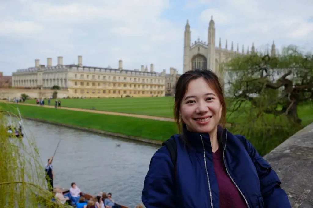 一位扎起头发、穿藏青色外套的女性微笑面对镜头,身后是英国剑桥的学院建筑,脚下是康河,河上有撑篙的平底船。