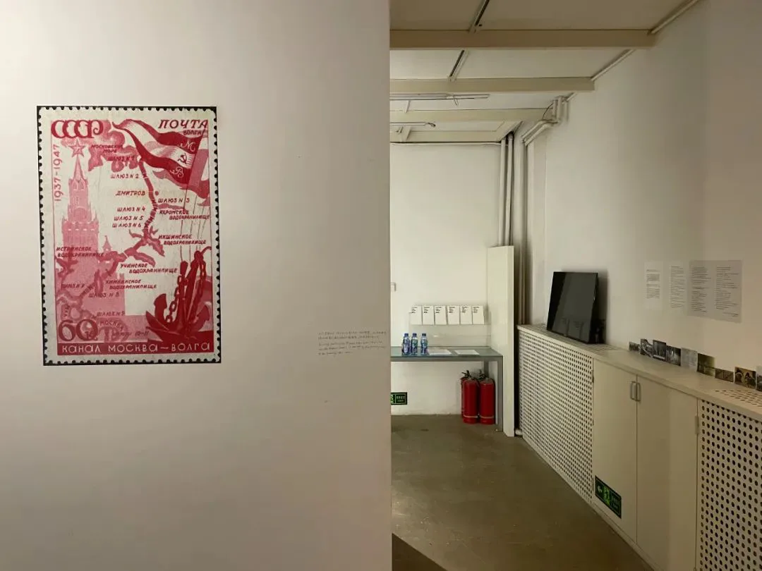 展墙左侧放大展示一张苏联红色主题邮票,印有莫斯科克里姆林宫图案、运河路线图和文字「1937-1947 运河莫斯科—伏尔加」;右侧是展厅内部的白墙走廊,墙上挂着一台平板电视和说明文字,柜台上摆放消防器材。
